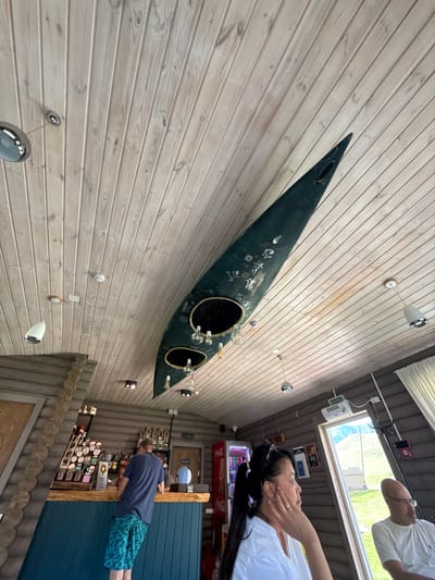 In Kupchegen, Russia, an interior scene depicts a bar area with a unique ceiling. The ceiling is constructed from light-colored wooden planks, running diagonally across the frame. Suspended from the ceiling is a dark green, elongated object resembling a canoe or kayak, cut lengthwise and mounted as a decorative element. It has two large circular openings, and several pendant lights with exposed bulbs hang from within.

Below, a man in a blue t-shirt and patterned shorts stands at a counter, appearing to order or interact with someone behind it. The bar itself has a teal-colored front and a wooden countertop. Shelves behind the counter display bottles and various food items.

To the right of the frame, two people are seated, facing away from the bar. One is a woman with dark hair pulled back, wearing a white shirt, and resting her chin on her hand. Beside her is a man with glasses and a bald head, also wearing a white shirt.

Further back, a Coca-Cola branded refrigerator is visible. A large window on the right side of the image looks out onto a grassy outdoor landscape with hills in the distance. The overall ambiance suggests a casual establishment, possibly a cafe or restaurant.