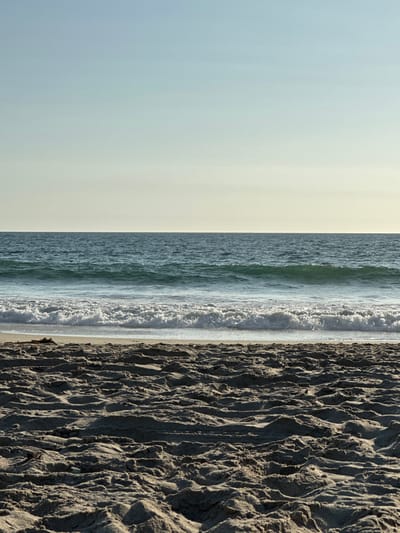 This image depicts a beach scene at Manhattan Beach, United States, captured from a low vantage point. The foreground is dominated by uneven, light brown sand with visible footprints and small ridges, indicating recent pedestrian activity. The midground features the Pacific Ocean, with turquoise-blue water exhibiting several parallel waves. The most prominent wave is breaking, forming a white foamy crest across the width of the frame, approximately mid-height. Beyond this, the water's surface shows gentle ripples and a darker blue hue. The background consists of a clear sky, transitioning from a pale yellow-white near the horizon to a faint light blue at the top of the frame. The horizon line between the ocean and sky is flat and uninterrupted. No people, animals, or man-made structures are visible within the frame. The scene is illuminated by natural daylight, casting subtle shadows on the sand, suggesting a sun angle from the left or upper left.