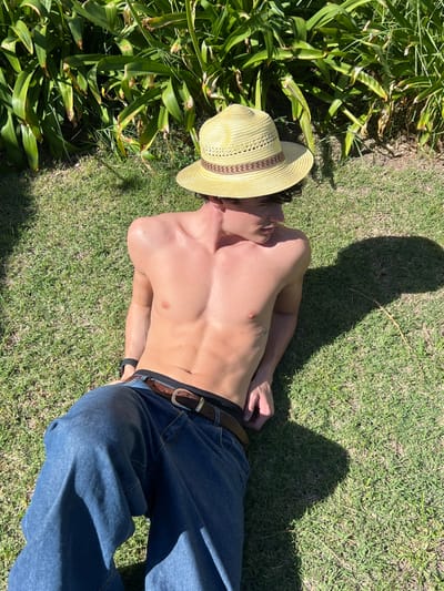 A young male is depicted lying on green grass outdoors in Quilmes Oeste, Argentina, under direct sunlight. He is shirtless, displaying a lean physique with visible abdominal muscles. He wears a light-colored straw hat featuring a brown woven band, blue denim jeans, and a brown belt with a silver-colored buckle. Black underwear is partially visible above the waistband of his jeans. A dark wristband is present on his left wrist. The male's head is turned to his right, and his gaze is directed downwards. His left arm is bent, while his right arm rests along his side on the grass. The background consists of dense green foliage, and sunlight casts distinct shadows on the grass to the right of the subject.