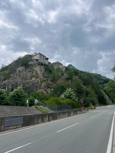 The image depicts a roadside view of a steep, rocky hillside on an overcast day. A paved road with white lane markings curves to the right, following the contour of the hill. A concrete retaining wall runs along the left side of the road, with a metal grate visible on its surface. Above the wall, a metal fence with green railing encloses a grassy area and a staircase leading up the slope. A light gray, multi-story building with balconies and a rounded tower stands atop the rocky outcrop, surrounded by lush green trees. The sky is filled with heavy, gray clouds. The scene is devoid of any people or vehicles. This location is identified as being in the city of Krichim, Bulgaria.