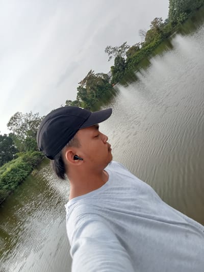 The image shows a person wearing a black baseball cap and a light gray t-shirt, looking to the right with their eyes closed. They have an earbud in their right ear. The person is standing on a platform next to a body of water, which appears to be a lake or a river. The water has ripples on its surface and reflects the overcast sky and the surrounding greenery. Lush vegetation, including trees and bushes, lines the edge of the water. The background shows more trees and foliage, with a cloudy sky overhead. The location is identified as Kualu, Indonesia, suggesting an outdoor setting within or near the city. The scene depicts a moment of quiet contemplation or relaxation by the water.