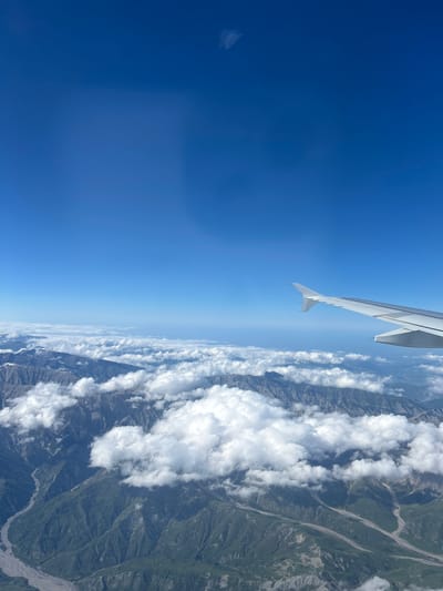 The image presents a striking aerial view, rotated 90 degrees clockwise, giving the impression of looking from an airplane flying with a significant bank. A vast, clear deep blue sky occupies the left side of the frame. Extending diagonally from the upper-middle towards the lower-left is the clean white wing of an airplane, establishing the perspective from within the aircraft.
To the right, a dramatic mountainous landscape unfolds beneath scattered white cumulus clouds. The rugged mountains feature deep valleys and varied topography, with visible rocky outcrops and patches of green vegetation. Lighter, winding lines are prominent within the valleys, suggesting rivers, riverbeds, or geological formations carved by water. The bright sunlight indicates it is daytime with excellent visibility, highlighting the textures and contours of the terrain. The scene depicts a high-altitude flight over a majestic mountain range, consistent with the geological features found in the Ismailli District of Azerbaijan. No text is visible in the image.