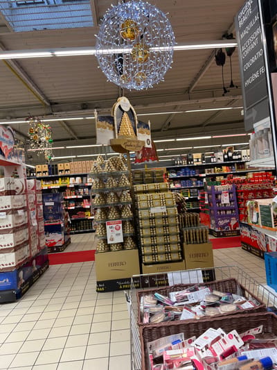 The image displays the interior of a hypermarket or supermarket located in Hérouville-Saint-Clair, France, characterized by holiday-themed displays. A large spherical light fixture composed of numerous white LED lights, containing three larger golden orbs, hangs from the industrial-style ceiling which features exposed utilities and fluorescent lighting strips, as well as a visible skylight. To the left, a smaller decorative wire structure adorned with colorful baubles is present.
The central area is dominated by an extensive Ferrero Rocher chocolate promotion. This includes a tall display stand with a graphic of a chocolate pyramid and the "Ferrero Rocher" brand name, supported by multiple tiered displays of individually wrapped Ferrero Rocher chocolates arranged in pyramid shapes on golden bases. Adjacent to these are large stacks of golden-packaged Ferrero Rocher boxes, with some visible pricing such as "12,99€."
On the left side, stacked cardboard boxes of Lindt chocolates are visible, and further into the background, a "Kinder" branded display is partially seen. The right side of the frame features an overhead sign listing product categories in French: "HYGIÈNE BEAUTÉ" (Hygiene Beauty), "MAQUILLAGE" (Makeup), "ACCESSOIRE COIFFANT" (Hair Accessory), "COLORATIO" (Hair Dye), and "HYGIÈNE FE" (Female Hygiene). Below and behind this sign, shelves are stocked with various beauty and personal care products. Further back, shelves containing bottles of wine and other general merchandise are visible. In the immediate foreground on the right, a wire shopping basket is filled with various packaged cosmetics, including compacts and lip products.
The floor throughout the visible area is a light-colored tiled surface with a grid pattern. A red carpet strip runs down one of the main aisles in the midground. No individuals are discernible within the frame.