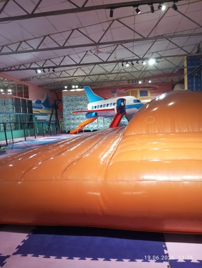 The image depicts an indoor play area, possibly an amusement park or a children's entertainment center, located in Kyzyl-Kyya, Kyrgyzstan. In the foreground, a large, inflatable orange structure dominates the scene, with a textured, rippled surface. Behind this structure, an artificial airplane serves as a play attraction, complete with a blue tail and white fuselage, and a red slide emerging from its side. The airplane is positioned against a colorful, patterned backdrop featuring abstract shapes and lines. The environment includes a ceiling with exposed metal beams and lighting fixtures, and the floor is covered with interlocking puzzle mats in purple and pink. The overall impression is a vibrant and playful setting designed for children's activities.