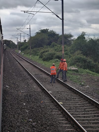 The image depicts a view of railway tracks near Khandwa, India, under a cloudy sky. Three individuals are present on the tracks. Two of them are wearing orange high-visibility vests and caps, with one of them appearing to be speaking. The third person is wearing a similar orange vest with "CENTRAL RAILWAY" printed on the back. They are standing on the ballast of the railway tracks, which are flanked by dense green foliage on the right. A train is partially visible on the left side of the image, suggesting it is either stopped or moving slowly. Overhead power lines for the electric train are visible stretching across the frame, supported by poles. The ground around the tracks is covered in dark grey ballast with some scattered litter. One pole has a yellow marker with black numbers "5658" on it. In the middle ground, a person carrying a large white sack is walking along the tracks.