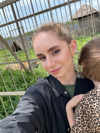 The image, taken in Timofeevka, Russia, captures a woman and a child visiting an animal enclosure, likely at a zoo or animal park. The scene is outdoors, illuminated by bright daylight under a clear blue sky.
In the foreground, a woman with light-colored hair, pulled back, is visible in a close-up, smiling slightly towards the camera. She has noticeable eye makeup, including eyeliner and defined eyebrows, and wears a small gold heart-shaped earring in her left ear. She is dressed in a dark (possibly black) jacket over a green top.
Partially visible on the right side, close to the woman, is a young child. The child has light brown hair and is wearing a top with a distinct leopard print pattern. A small hand is visible, resting on the woman's arm.
Behind the two individuals, and separated by robust metal bars of an enclosure, a tiger is visible. The large, striped cat's head and upper body are seen amidst green grass and vegetation. The tiger appears to be looking in the direction of the camera or the people. In the background within the enclosure, a wooden structure, possibly a shelter or den, is also discernible. The metal fencing signifies a safe viewing barrier between the visitors and the wild animal. The atmosphere appears pleasant and bright, consistent with a daytime visit to an animal attraction.