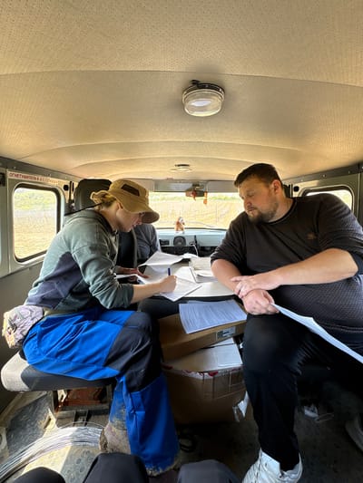 Two individuals are inside a vehicle, likely a van or truck, in Zaprudnoe, Ukraine. One person, wearing a black patterned shirt and dark pants, is standing and holding papers. The second person, seated and wearing a khaki cap, green shirt, and blue work pants, is also holding papers and a pen. Both individuals appear to be examining the documents. Additional papers and a cardboard box are visible on the dashboard. The vehicle's interior has light-colored perforated upholstery. Outside the window is a rural landscape with trees and fields.