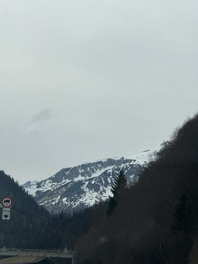A landscape photograph shows a snow-covered mountain next to dark trees and an overcast sky. A road sign with an arrow is in the lower right corner. The sign has the word "Anfang" and a symbol for maximum vehicle height of 3.5 tons. This image was taken in the vicinity of Rothenwand, Austria.