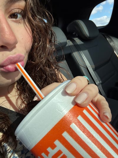 A woman with brown curly hair sips from an orange-striped Whataburger cup using a straw. She is inside a car; black leather seats are visible in the background, along with a glimpse of a blue sky. Her fingernails are painted a neutral color. A turquoise ring adorns her finger. The image focuses on her face and the cup.