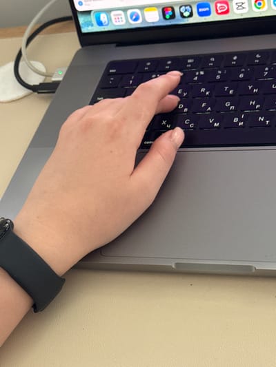 A close-up, elevated shot shows a person's left hand and wrist, adorned with a black smartwatch, resting on the keyboard of a grey laptop. The hand is positioned as if typing, with fingers poised over the keys. The laptop displays several application icons at the top, including Zoom, suggesting a digital environment. In the background, a white cord is plugged into the laptop, with a small green light indicating it is active. The scene appears to be set on a light-colored, textured surface, possibly a desk. The image captures a moment of digital interaction, with the focus on the user's hand and the technology it engages with. The context of Troitske, Ukraine, is not visually present in the image itself, as the scene is a generic representation of someone using a laptop.