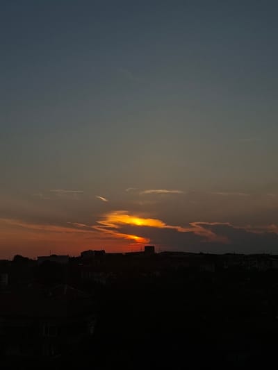 This image captures a sunset over what is identified as the city of Galabovo, Bulgaria. The upper sky is a dark, muted blue-grey, transitioning into warm hues of orange, red, and intense yellow closer to the horizon. The setting sun, visible as a brilliant yellow-orange glow, is partially obscured by elongated, horizontal clouds and some wispy streaks, which are illuminated by the sun's light. Below the horizon, the landscape is rendered in deep silhouette, comprising an urban or suburban area. Numerous buildings of various sizes and shapes, along with dense trees and foliage, form a dark, undulating skyline. Taller structures, possibly industrial or multi-story residential buildings, are discernible as dark masses on the horizon, particularly towards the left and center. The foreground is almost entirely black, consisting of further obscured urban elements and vegetation, lacking specific detail. No individuals or specific human activities are visible within the frame.