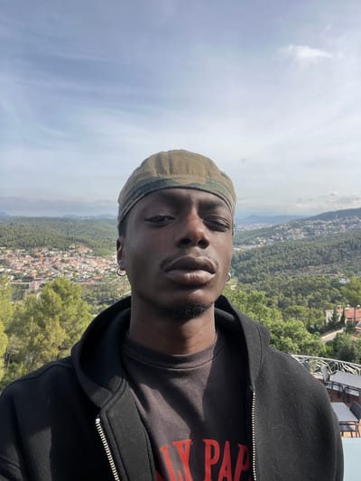 A medium shot portrait of a young Black man wearing a camouflage baseball cap, a black zip-up hoodie over a dark t-shirt, and a hoop earring in his left ear. He is standing outdoors with a city and mountainous landscape in the background. The city appears to be Can Güell, Spain, with buildings nestled into green hillsides under a partly cloudy sky. The man is looking directly at the camera with a neutral expression, his left eye squinted. The lighting suggests it is daytime.
