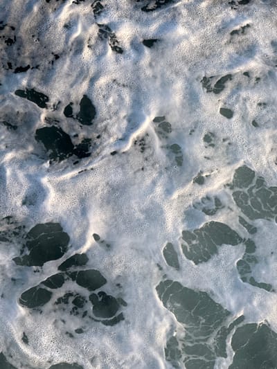 The image presents an overhead perspective of turbulent seawater off the coast of Kato Karioutes, Greece. The scene is characterized by a high volume of white sea foam, composed of numerous small bubbles, forming irregular masses and streaks across the surface. These foamy areas contrast sharply with darker, exposed sections of water, which exhibit subtle currents and variations in tone. No human figures, animals, or specific man-made objects are visible within the frame. The overall environment is marine, depicting active water movement characteristic of waves breaking or strong currents.
