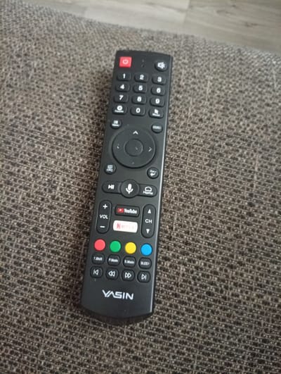 A black YASIN brand TV remote control is lying on a textured gray fabric surface. The remote has a power button at the top, followed by a numeric keypad (0-9), and directional buttons surrounding a central selection button. Below the numeric keypad are buttons labeled "Favorite", "Menu", and "Source". Further down are buttons for "EPG", "Mute", a microphone icon, and "Home". There are also volume up and down buttons, and buttons for YouTube and Netflix. The lower section of the remote has colored buttons (red, green, yellow, blue) and playback controls. The image is taken from a slightly elevated angle, focusing on the remote. The background shows a blurred gray and brown textured surface, likely upholstery. The location of Semetey, Kyrgyzstan is not discernible from the image itself.