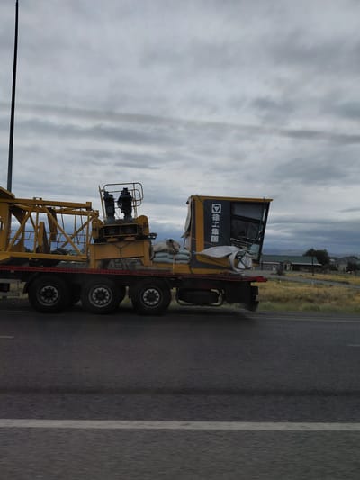 A yellow crane is being transported on a flatbed truck on a highway. The truck has multiple wheels and the crane appears to be disassembled or in a transport configuration. There are bags of sand on the back of the truck. The background shows a dry, grassy landscape with a few houses and buildings. The sky is cloudy. The scene is likely in the city of Usharal, Kazakhstan, given the context.