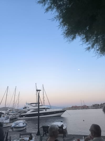 A serene scene unfolds at dusk in a Greek harbor, possibly Chalki. The sky is a gradient of soft blue and pale pink, with a faint moon visible. In the foreground, people are seated at tables, some facing away, suggesting a relaxed evening gathering. The harbor is filled with various boats, including several sailboats with tall masts and a sleek, modern yacht docked prominently. Smaller vessels, like a blue and white dinghy, are moored nearby. Beyond the boats, a picturesque coastal town with red-roofed buildings lines the waterfront. The overall atmosphere is peaceful and tranquil, indicative of a calm evening in a Mediterranean setting. The time of day is clearly sunset or twilight, with soft lighting casting a gentle glow.