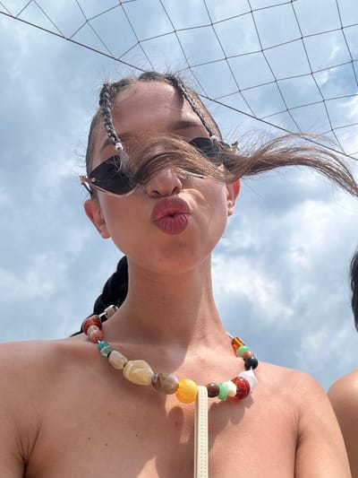 This media file shows a close-up, rotated shot of a young person wearing sunglasses and a beaded necklace. The person's lips are puckered, giving the impression of blowing a kiss. They have dark hair styled in braids, with a few strands loose around their face. The background is a cloudy sky, with a net, possibly from a volleyball court, visible on the left side of the frame. The lighting suggests it is daytime, possibly late afternoon or early evening, with soft, diffused light. The setting appears to be outdoors, in a recreational area near Ravadinovo, Bulgaria, indicated by the volleyball net and the bright, open sky. The overall mood is playful and relaxed. There is no visible text in the image.
