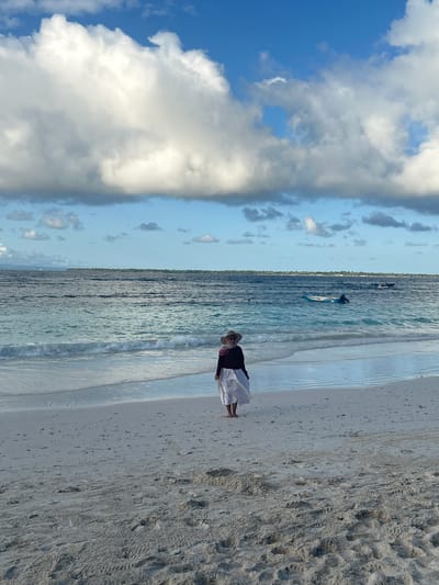 A woman wearing a straw hat, dark top, and white skirt stands on a sandy beach facing the ocean in A R A, Indonesia. The ocean water is a turquoise blue with gentle waves. In the distance, several boats are visible on the water, and a long strip of land is on the horizon. The sky is blue with scattered white and gray clouds. The sand is light in color and shows imprints of footprints.