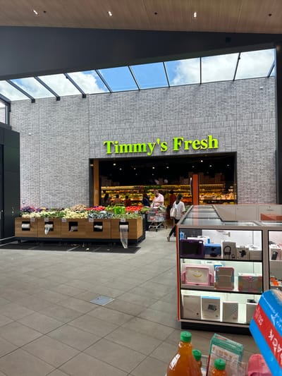This image captures the entrance of "Timmy's Fresh," a grocery or fresh food market located within a modern shopping complex in Clyde North, Australia. The store's name, "Timmy's Fresh," is displayed in bright green lettering against a light grey brick facade.

The setting is bright and spacious, characterized by a high ceiling featuring multiple large skylight windows that reveal a clear blue sky with white clouds, indicating it is daytime. The floor is tiled with large, light-colored square tiles. In the foreground and extending towards the store entrance, a vibrant display of fresh produce, including various fruits and vegetables, is neatly arranged on wooden shelving units and in bins.

Several individuals are visible, engaged in shopping activities. One person, wearing a light-colored shirt, is seen pushing a shopping cart near the entrance, either entering or exiting the store. Other shoppers are browsing the produce sections within "Timmy's Fresh." Adjacent to the store entrance, a display cabinet showcases various merchandise, including what appear to be tablet cases and other electronic accessories. In the immediate foreground, partially visible, are a plastic bottle of orange juice and a blue multi-pack item with the word "PACK" printed on it. The scene depicts a bustling retail environment with shoppers purchasing groceries and other goods.