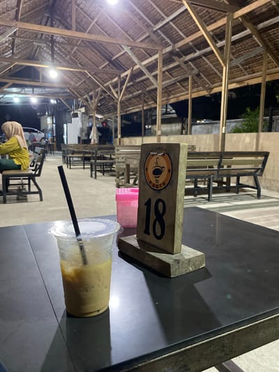 The image depicts an outdoor restaurant setting in Gampongaceh, Indonesia, at night. The roof structure is made of wood beams and thatch. Numerous metal tables and chairs are arranged in rows. In the foreground, a black metal table holds a plastic cup with a straw, containing a yellow beverage. A wooden table marker with the number "18" and a logo is also present. In the background, a person wearing a yellow top and hijab is near a table. The overall atmosphere is illuminated by artificial lights.