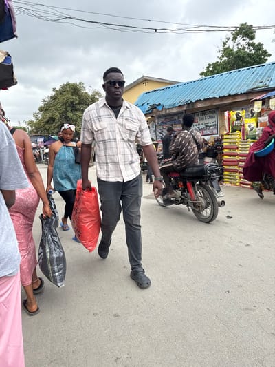 The image depicts a bustling street scene in Ajah, Nigeria, characterized by pedestrian and motorcycle traffic alongside market stalls.
In the foreground, a Black male, estimated to be between 25-35 years old, walks towards the left of the frame. He wears a light-colored plaid short-sleeve button-up shirt over a dark undershirt, dark jeans, and dark closed-toe sandals. He has dark sunglasses on and a short, dark haircut. He carries a large, full red plastic bag in his right hand and a smaller black plastic bag in his left hand. To his immediate left, partially obscured, are other pedestrians, including a female in a pink patterned garment carrying a black plastic bag and another in a light grey t-shirt.
In the midground, to the left of the central figure, a Black female, approximately 20-30 years old, walks away from the viewer. She wears a blue halter-neck top, dark leggings, and a white patterned headband, holding a small white water bottle. To the right of the central figure, a Black male, approximately 20-30 years old, sits on a red motorcycle, facing away from the viewer. He wears a dark patterned long-sleeved shirt.
The background features market structures. A building with a light blue corrugated metal roof runs along the right side of the street. Underneath, signs are visible, one advertising "JAMIE COLLEGE & CULTURAL ORGANIZATION" and educational degrees (B.Ed, B.A, Ed. Local & Int'l) with a phone number (08064974999). Another sign advertises "QUALITALL GOLD" with images of animals, positioned above stacks of yellow and green bags (likely feed or produce). A person in a red robe carrying a light blue basin is visible on the far right. Further left in the background, trees and more distant market activity are present. The ground is a light-colored concrete or paved surface, showing some wear. Overhead, power lines crisscross against a cloudy, overcast sky.