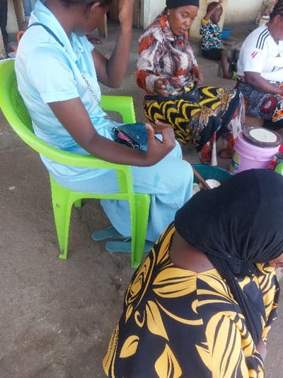 The image depicts an outdoor or semi-outdoor scene in Kimamba, Tanzania, featuring four adults and one child engaged in various activities on a light gray concrete or dirt surface. In the foreground, the back and left shoulder of an individual wearing a black headscarf and a black garment with yellow leaf patterns are visible. To the left, a young adult female is seated on a bright green plastic chair, wearing a light blue short-sleeved collared shirt and matching trousers. Her right hand is raised near her temple, and her left hand holds a dark blue rectangular purse with a silver chain strap. Light blue flip-flops are visible on the ground near her feet.
In the mid-ground center, an adult female is seated directly on the ground. She wears a dark head covering and a long-sleeved shirt with orange, brown, and black abstract patterns, over a yellow and black patterned skirt. She is actively engaged in food preparation, using both hands to manipulate a light-colored dough-like substance within a white bowl situated inside a larger green plastic basin.
To the right of this woman, another adult female is partially visible, also seated on the ground. She wears a white short-sleeved t-shirt with a red logo on the chest and a patterned skirt. In front of her, a pink plastic container holds a flat, round, white disc, consistent with an uncooked flatbread or dough. In the background, a small child wearing a blue and white patterned long-sleeved shirt is seated or crouching on a raised concrete step or platform. A light-colored wall forms the immediate background.