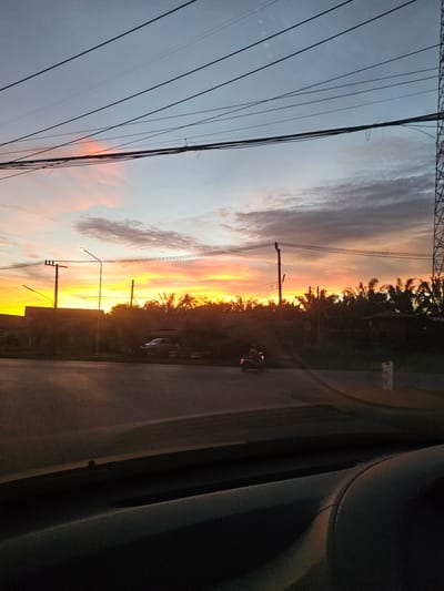 This media file is a photograph taken at sunset in Khlong Thom, Thailand. The scene depicts a vibrant sunset sky with hues of orange, yellow, and pink, casting a warm glow over the landscape. Several utility poles and power lines are prominent in the foreground, stretching diagonally across the frame. In the middle ground, a road is visible, with a motorcycle and what appears to be a truck parked or driving on it. The background is filled with silhouettes of trees and vegetation, suggesting a rural or suburban setting. The overall mood is serene and picturesque, capturing the beauty of a tropical sunset. The time of day is clearly indicated by the sunset, and the weather appears clear, allowing for the unobstructed display of the colorful sky. No visible text is present in the image.
