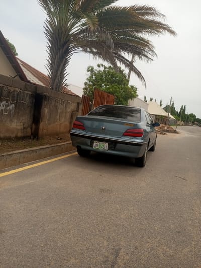 A teal-colored Peugeot 406 sedan is parked on the right side of an asphalt road, facing away from the camera. The car has a license plate that reads "JMT-842AH". To the left of the road is a concrete wall with some graffiti, and behind the wall are houses with corrugated metal roofs and a palm tree. The sky is overcast. The scene appears to be a residential area in Baccure Kofare, Nigeria.
