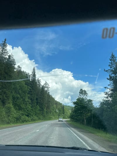 A white car drives down a paved road through a forest under a bright blue sky with fluffy white clouds. The road is lined with trees and vegetation. The image appears to have been taken from inside a vehicle, possibly through the windshield. There is a dark dashboard visible at the bottom of the frame. In the upper right corner of the image, there is a blurred marking that appears to be "00-" on the windshield. The specific location in the city of Усмангали, Russia is not identifiable from the visual information provided.