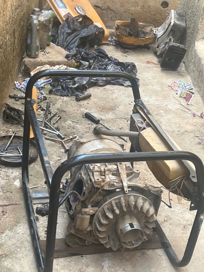The scene depicts a partially disassembled portable generator, likely undergoing repair. It is situated on a concrete surface in what appears to be an outdoor or semi-outdoor location, possibly in Unguwar Fatika, Nigeria. The generator's frame is made of black metal tubing. Key components, such as the engine, alternator, and carburetor, are visible and appear to be covered in dust and grime. Several tools, including wrenches and a screwdriver, are scattered around the generator, suggesting ongoing maintenance. There is also a collection of other items nearby, including black plastic bags, empty plastic bottles, and a yellow fuel tank. The overall environment is somewhat cluttered. There is no evidence of people in the image.