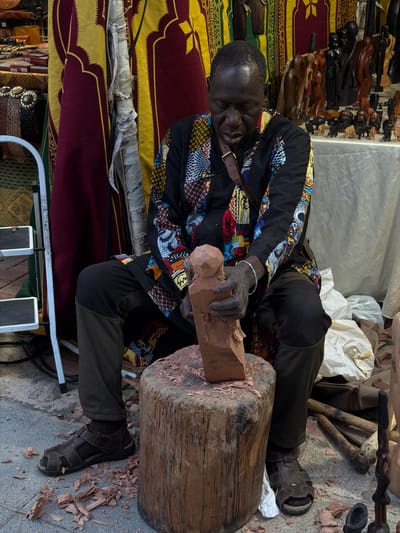 A craftsman is seated on the ground in what appears to be a market setting in Burjassot, Spain. He is positioned next to a wooden stump used as a workbench, where he is carving a piece of wood. He is wearing a patterned shirt, dark pants, and sandals. The ground around him is covered in wood shavings. Behind him, there is a display of other carved wooden objects. The location appears to be an open-air marketplace.