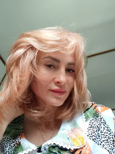 A medium close-up frontal shot shows a light-skinned woman with blonde, shoulder-length hair and pink lipstick. She is looking slightly off to her right, with her left arm and hand resting on the left side of her neck. She is wearing a white, green, orange, and black patterned shirt with a lace trim at the collar. The background is a muted green wall with a wooden beam. The image was taken in Vytegra, Russia.