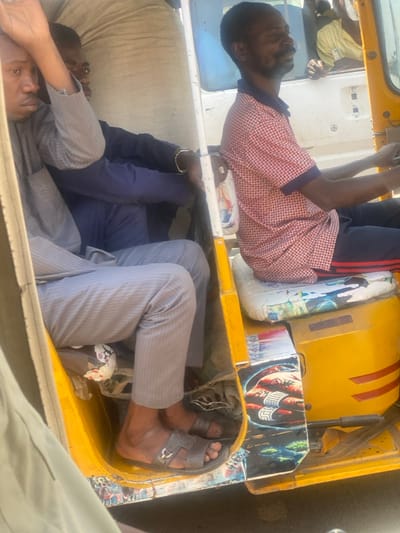 Within the city of Bachirawa, Nigeria, an image captures the interior and immediate surroundings of a yellow, three-wheeled auto-rickshaw during daylight hours. The vehicle contains three adult male occupants and cargo.
The driver, a dark-skinned male with a beard and short dark hair, is seated on the right, wearing a red and white checkered short-sleeved collared shirt with blue trim and dark pants. He is positioned at the controls, looking to his right, out of frame. His seat cushion is white and patterned, with a religious image of a woman visible on the backrest.
Behind the driver, a dark-skinned male passenger is seated to the left, wearing a light grey striped long-sleeved shirt and matching pants, with grey sandals on his feet. His left arm is raised and bent, resting against an unseen upper frame or cargo. His gaze is directed forward. Discarded white and red material is visible on the floor near his feet.
A second dark-skinned male passenger is partially visible, seated behind the first passenger and to his left. He wears a dark blue long-sleeved shirt and dark blue pants, with a gold-colored bracelet on his left wrist. Much of his upper body is obscured by a large, light-colored sack positioned as cargo in the rear compartment.
The auto-rickshaw's interior features decorative elements, including a decal of a red fist wearing rings on the lower right-side panel. A white vehicle with a partially visible person in a yellow shirt is present in the background, seen through the driver's side window.