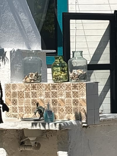 The image shows a close-up of a decorative shelf unit located outdoors, likely in Varvara, Bulgaria. The shelf is topped with decorative tiles and features three glass jars filled with seashells. A sink with a faucet and a bottle of blue liquid soap are also visible on the shelf, suggesting this might be an outdoor wash area. The background includes a window with blinds and a curtain, casting shadows across the scene. The overall impression is one of a quaint, possibly rustic, outdoor setting.