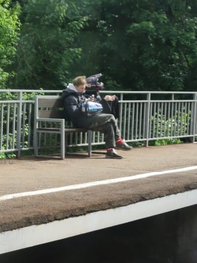 The image shows two people sitting on a bench next to a metal fence on a train platform in Ashukino, Russia. One person, on the left, is wearing a dark jacket, camouflage pants, red socks, and dark shoes, and is looking down at something in their hands. The other person, on the right, is wearing a black jacket, a helmet, and is also looking down at something in their hands. The background is a green, leafy area. The platform edge is visible in the foreground.
