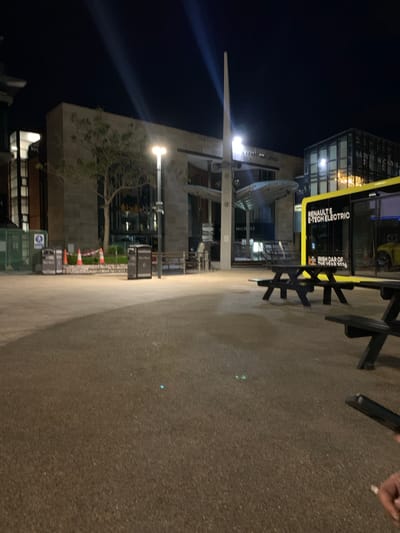The image depicts a nighttime scene on a paved pedestrian area in Dublin 16, Ireland. A modern building with glass and concrete architecture is visible, featuring signage with "RENAULT & E-TECH ELECTRIC" and "IRISH CAR OF THE YEAR 2026" printed on it. There is outdoor seating with black picnic-style tables and benches. Construction cones are lined up along the side of the building. A tall metal structure resembling a spire is partially visible. A person's hand holding an object is in the top right corner of the image.