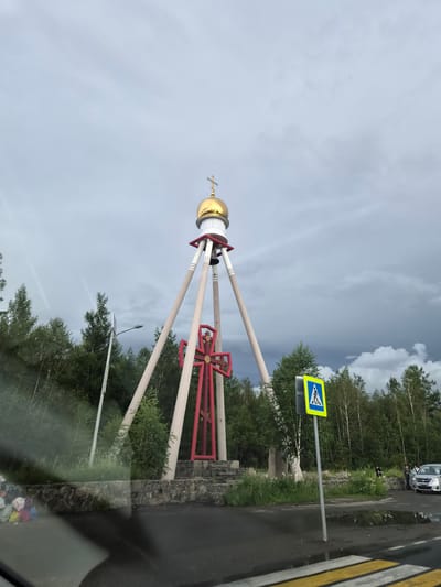 The image depicts a large red cross with a golden dome on top of a white structure, supported by three light-colored poles. The structure is situated in an outdoor environment with a treeline of green coniferous trees in the background. A pedestrian crossing sign is visible to the right of the cross. The sky is cloudy and overcast. In the foreground, there is a wet asphalt road with yellow and white stripes indicating a crosswalk. To the left, near a stone wall, several colorful bags are piled up. A silver car is parked on the right side of the road, with two people standing near it. The location is identified as Verkh-Chita, Russia.