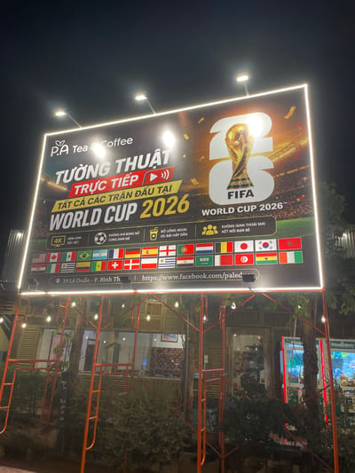 A nighttime view shows a large, illuminated advertisement for the FIFA World Cup 2026 at P.A Tea & Coffee. The sign is in Vietnamese, announcing the live broadcast of all matches at the World Cup. It features the FIFA logo, the World Cup trophy, and a list of participating countries' flags. The sign is located at 39 Lê Duẩn Street in P. Bình Th, Vietnam. The surrounding environment includes outdoor seating, overhead lighting, and some foliage.