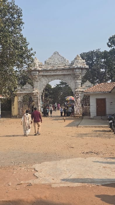 The image depicts the entrance to a complex in Katwaldih, India, under clear daylight conditions. A prominent, ornate white stone archway with intricate carvings forms the main gateway, featuring a large central arch flanked by two smaller arches. The ground in the foreground is unpaved, consisting of reddish-brown dirt and gravel, with a patch of lighter grey concrete visible on the right.

In the immediate foreground, two individuals walk away from the camera towards the archway. The person on the left wears a light-colored, possibly white or cream, traditional Indian outfit, while the person on the right wears a maroon long-sleeved shirt and light brown trousers. A small, light brown dog walks ahead of them, slightly to the right of the center.

Beyond the main archway, a crowd of people is visible entering or moving within the complex. To the immediate right of the central arch, a vendor stall is set up, identified by a vibrant, multi-colored striped umbrella (red, yellow, blue). Further to the right of the archway stands a small, white, single-story building with a terracotta-tiled hip roof and a brown wooden door. A black motorcycle is parked near this building. On the left side, thick green foliage from trees is visible, with hints of a smaller gate structure. Trees with dense green leaves frame the upper left and right sides of the image and extend into the background beyond the archway. The sky above is clear and light blue. Shadows indicate a high sun position.