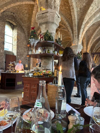 The image depicts an indoor dining or event setting within a large, vaulted stone hall in Cernay-la-Ville, France. The space features exposed light-colored stone walls, a high vaulted ceiling supported by numerous thick stone pillars, and an arched window visible on the far left.
Several pillars are adorned with multi-tiered shelves displaying festive decorations, including small evergreen trees with red bows, silver foliage, and multi-branched candelabras with white candles.
In the midground, a buffet is set up with two dark wooden counters. These counters hold an array of food items in silver serving dishes, chafing dishes, and tiered stands, including various salads and other dishes. A man in a white chef's jacket stands behind the left buffet counter, appearing to attend to the food. Another man, wearing a beige jacket, is partially obscured by a stone pillar near the center buffet. In the midground-right, two women are seen from behind, standing or moving near the buffet area; one wears a light grey textured vest, and the other wears a dark, possibly faux-fur, jacket.
The foreground shows a wooden dining table set with white plates, multiple wine glasses containing white wine, a clear glass bottle of Badoit mineral water, and a silver candlestick holding a lit white candle. A plate on the table contains seafood, specifically prawns and crab claws, accompanied by lemon wedges. A person's hand is visible on the left, near a plate, and a male diner is partially visible on the right, holding a glass. The scene is illuminated by ambient light and the flickering candle flame.