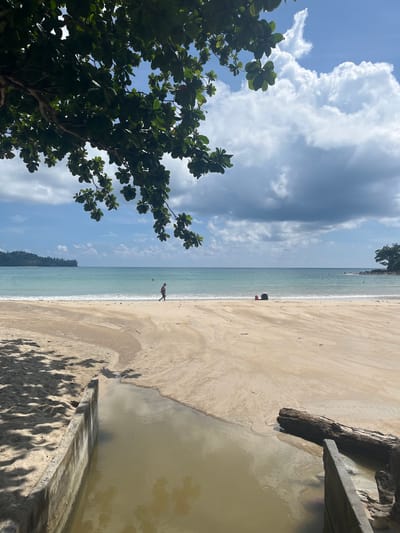 The image depicts a serene beach scene in Ban Bang Thao, Thailand, on a bright day with scattered clouds. In the foreground, a narrow channel of murky water runs from the left towards the sea, bordered by concrete structures. The sandy beach is wide and gently slopes towards the turquoise ocean. In the middle ground, a lone individual is walking along the shoreline, while further out, two red and black objects, possibly bags, rest on the sand. The background features a calm sea meeting a hazy horizon, with lush green hills visible in the distance on the left and a rocky outcrop with trees on the right. Overhanging branches of a large tree partially shade the scene from the top left.