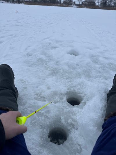The image captures a first-person perspective of an ice fishing scene on a frozen, snow-covered body of water. The dominant feature is the white, textured snow and ice that blankets the surface.
In the immediate foreground, the lower half of a person's body is visible. They are wearing dark, rugged boots and blue or dark-colored pants. Their right hand, clad in a dark glove or sleeve, firmly grips a bright yellow ice fishing rod. The rod has a small, red-colored tip and extends downwards towards one of the fishing holes.
In the midground, three dark, circular holes are visible in the ice, indicating where the ice has been drilled for fishing. Two of these holes are more prominent, closer to the viewer, while a third is slightly further back and to the left. The snow around these holes shows some disturbance and accumulation of ice shavings, suggesting recent activity. The holes appear dark and deep, contrasting sharply with the surrounding white snow.
The surface of the snow displays various textures, with some areas appearing smooth and others lumpy or crystalline, suggesting a mix of freshly fallen snow and possibly older, compacted ice and snow.
In the distant background, along the edge of the frozen expanse, there are bare trees and what appear to be some structures or houses, indicating that the location is not entirely remote. The sky, though not fully visible, seems to be overcast, contributing to the soft, diffused lighting and suggesting cold, wintery weather conditions suitable for ice fishing.
The overall impression is one of quiet, cold winter activity, focusing on the immersive experience of ice fishing.
