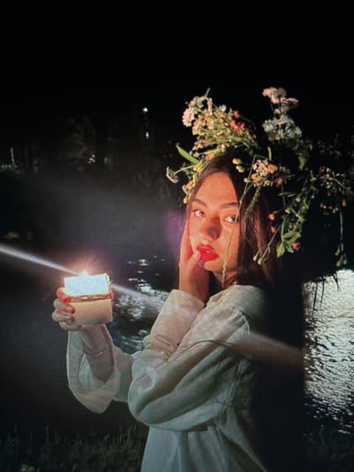 A young woman with a floral crown and bright red lipstick holds a lit candle in her left hand. She wears a white, long-sleeved garment. The background appears to be a body of water at night, with some vegetation visible. The woman's face is turned towards the camera, and she is touching her cheek with her right hand. The scene is dark and atmospheric, with the candle providing the primary light source. There is no specific indication of the location being Chotěšov, Czechia.