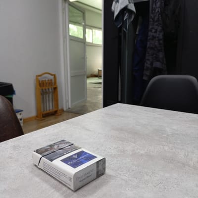 A pack of Parliament Night Blue cigarettes rests on a textured grey surface in the foreground. The pack is oriented diagonally, with the top flap open and slightly to the left. The branding "PARLIAMENT" is visible in white lettering on a dark blue background. In the background, a doorway leads into a brightly lit room with windows, suggesting an indoor environment. To the right of the doorway is a dark wardrobe with clothing hanging inside. Further to the left, a wooden rack with metal rods stands against a white wall. A dark chair is partially visible on the far right. The overall scene appears to be an interior setting, potentially an office or common area. The location in the city of Kinel, Russia, is not discernible from the visual elements present in the image.