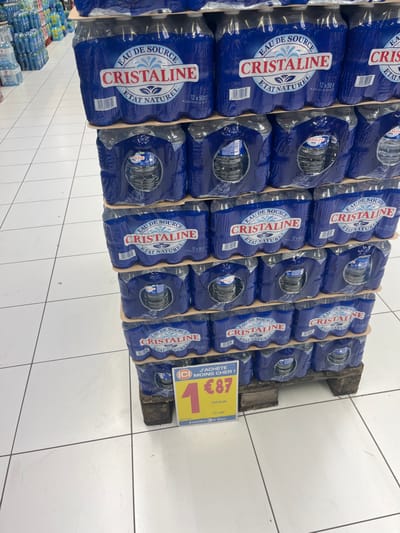 A stack of six packs of Cristaline brand bottled water is on a wooden pallet in a store in Saint-Brice-Courcelles, France. The bottles are arranged in six packs, with each pack containing six bottles. Each bottle has a blue label with the words "Cristaline Eau de Source Etat Naturel" on it, and the pallet is made of wood. A yellow sign in front of the pallet reads "J'achete moins cher!" with a number 1 and the price "€87" displayed prominently. The store floor is tiled in white and grey. The environment is clean and well-lit. There is no other person or object visible in the image.