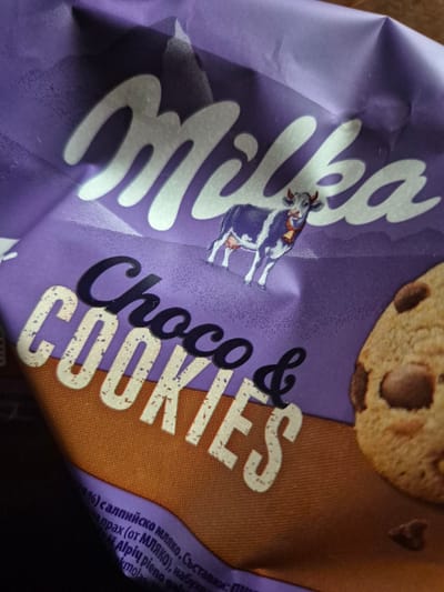 This is a close-up, top-down shot of a crumpled purple and brown Milka chocolate chip cookie package. The Milka logo, in white script, is prominently displayed across the top of the package. Beneath the logo, the words "Choco & COOKIES" are written in a combination of dark purple and off-white lettering. A cartoon cow, a brand mascot for Milka, is depicted in the upper-middle section of the image. The lower portion of the image shows a close-up of a chocolate chip cookie with visible chocolate chunks. Text in a foreign language is visible in the lower left corner, partially obscured by shadow. The image does not show any specific location within the city of Iskrets, Bulgaria.