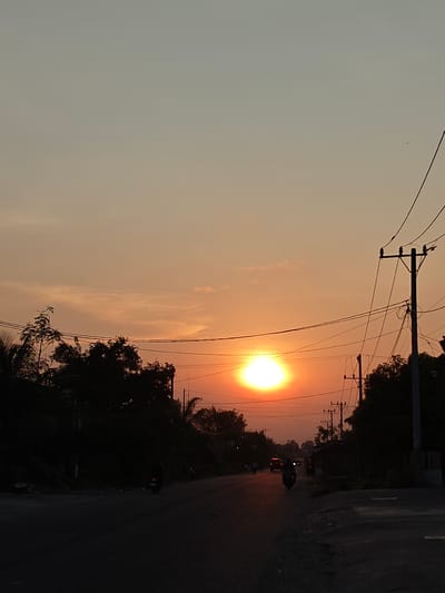 The scene depicts a sunset over a road in Blangcot Tunong, Indonesia. The sky transitions from a light gray-blue at the top to an intense orange and yellow glow at the horizon, where the circular, brilliant orange sun is visible, positioned slightly left of the image's vertical center. A dark asphalt road extends into the distance, appearing unlit in the foreground. Several vehicles are present on the road: at least three motorcycles, seen as dark silhouettes, with one motorcycle in the midground displaying a visible headlight, and one distant car showing illuminated red taillights. Dense, dark masses of trees and foliage flank both sides of the road. Multiple utility poles, laden with numerous wires, rise along the road's edge, primarily on the right, their dark forms silhouetted against the radiant sky.