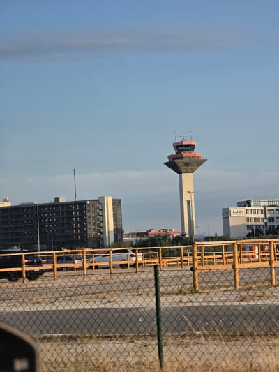 The image depicts an airport control tower in Walldorf, Germany, on a clear, sunny day with a few wispy clouds. The control tower, a prominent landmark, is tall and slender with a distinctive red and gray top section. To the left of the tower, a large, dark, multi-story building with many windows is visible. To the right of the tower, a modern white building displays the logo "LUG air cargo handling." In the foreground, a wooden fence runs horizontally across the frame, with a chain-link fence in front of it. Several vehicles are parked behind the wooden fence, including a black SUV and a white SUV. The ground appears to be a mix of gravel and pavement, with patches of dry grass. The overall scene suggests an operational airport environment.
