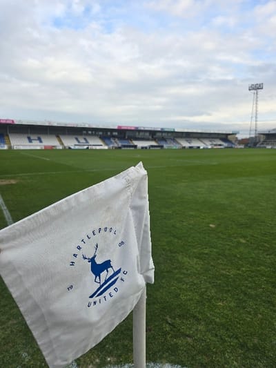 The image displays the interior of Victoria Park, the home stadium of Hartlepool United Football Club, located in Hartlepool, United Kingdom. In the foreground, a white fabric corner flag is prominently positioned at the intersection of white boundary lines on a green grass football pitch. The flag bears the blue crest of Hartlepool United FC, featuring a stag emblem, the club name, and the years "19" and "08". It is attached to a white pole. The midground consists of the manicured, empty green grass pitch. The background reveals the stadium's spectator stands, which feature multiple tiers of blue and white seating and various advertising hoardings. A tall floodlight tower stands on the right side of the stadium structure. The sky above is partially cloudy, indicating daylight conditions. The stadium appears unoccupied.