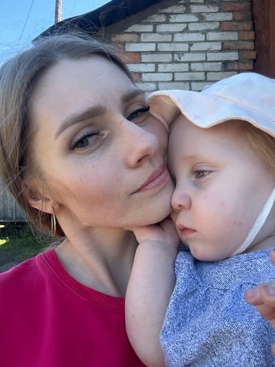 In Novoyeniseyka, Russia, a close-up selfie shows a woman and a baby. The woman, with light brown hair and light-colored eyes, is wearing a pink sweatshirt and a gold hoop earring. She is holding the baby close, with her cheek resting on the baby's head. The baby, with light blonde hair and fair skin, wears a pink hat with small hearts and a blue textured shirt. The background features a brick wall and a clear blue sky. The lighting suggests an outdoor setting during the daytime.